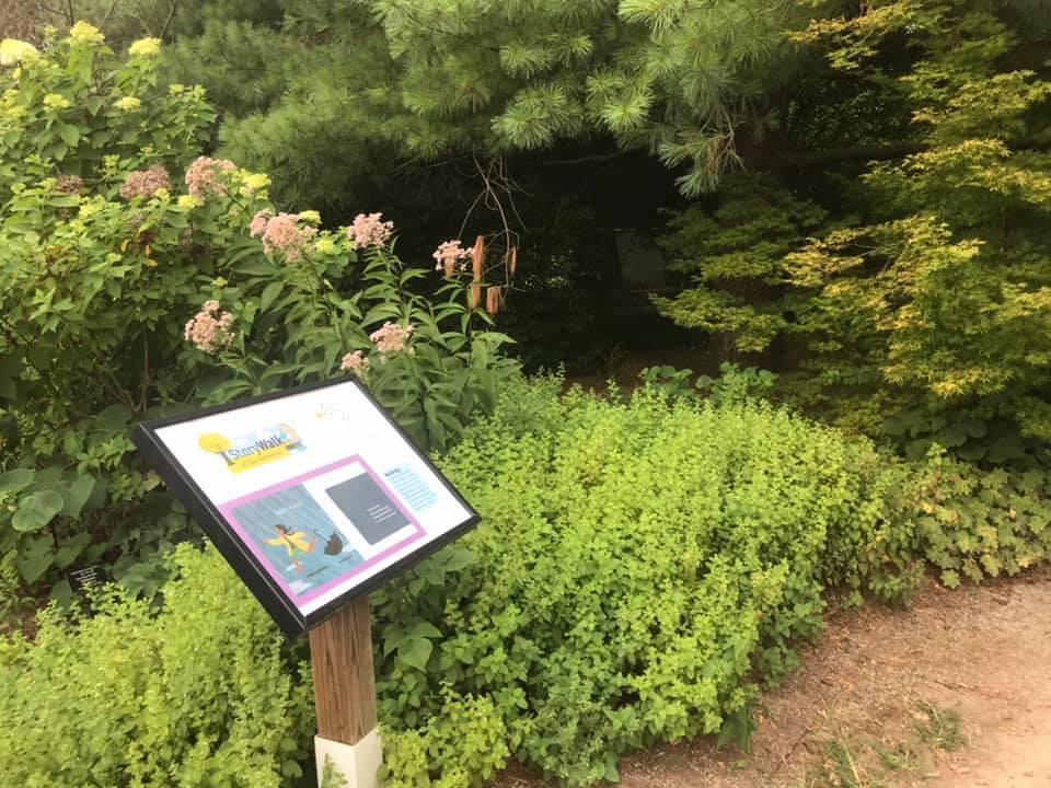 A StoryWalk sign surrounded by plants and trees in The Arboretum
